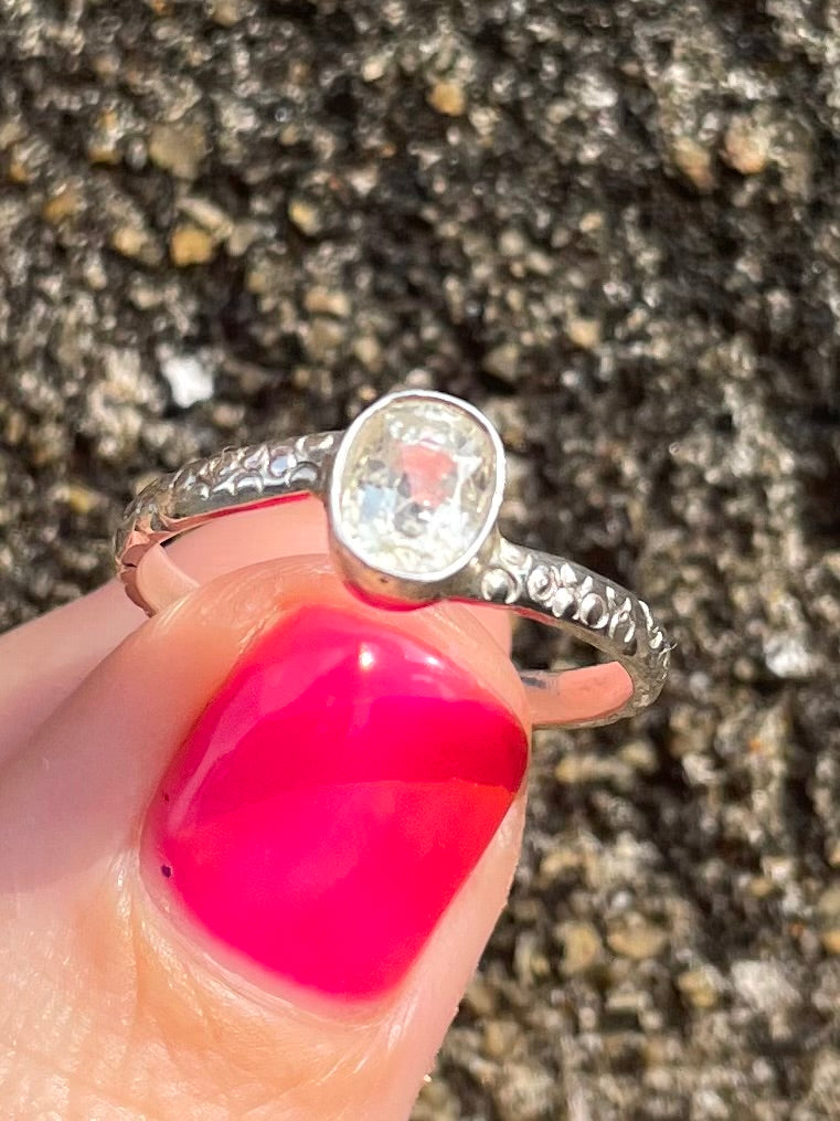 Silver ring with a large white sapphire held by a hand with pink nail polish against a textured background