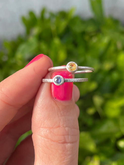 Two silver engraved rings with yellow and blue Sri Lankan Sapphires held by a hand against a blurred leafy background