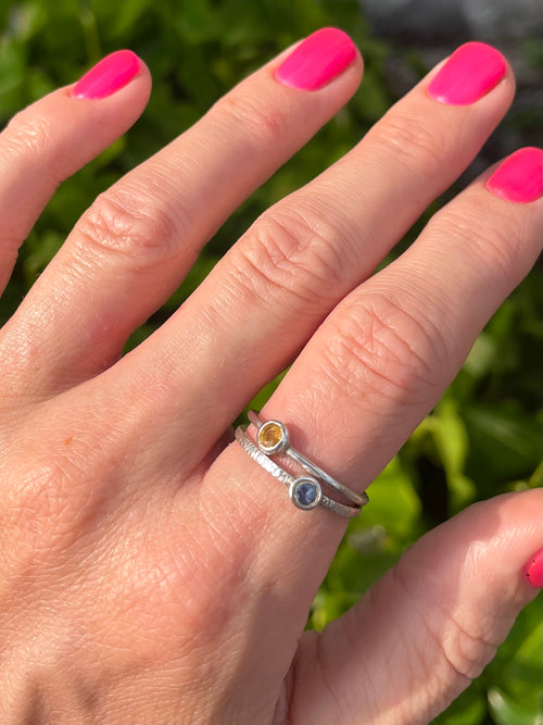 Hand with pink nail polish wearing two silver rings with blue and yellow sapphire on a green leafy background