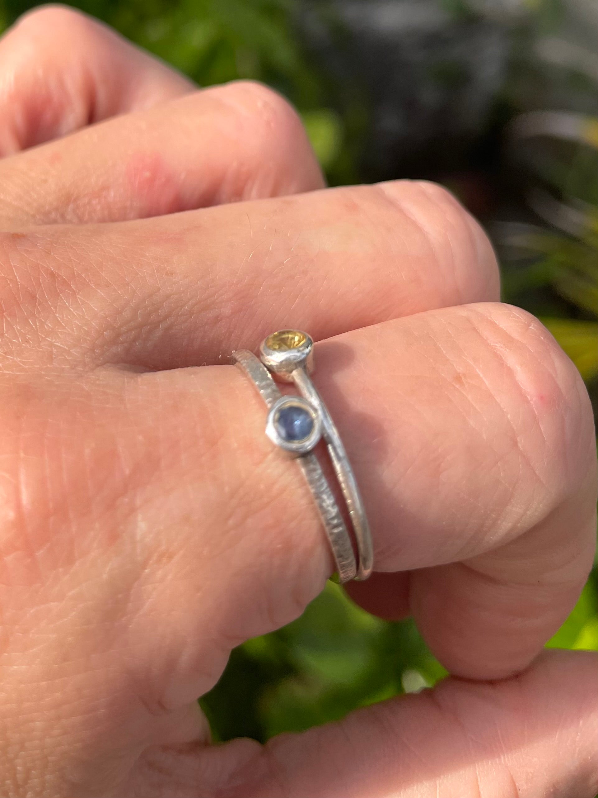 Close-up of a hand wearing two silver rings with Sri Lankan Sapphires against a blurred green background