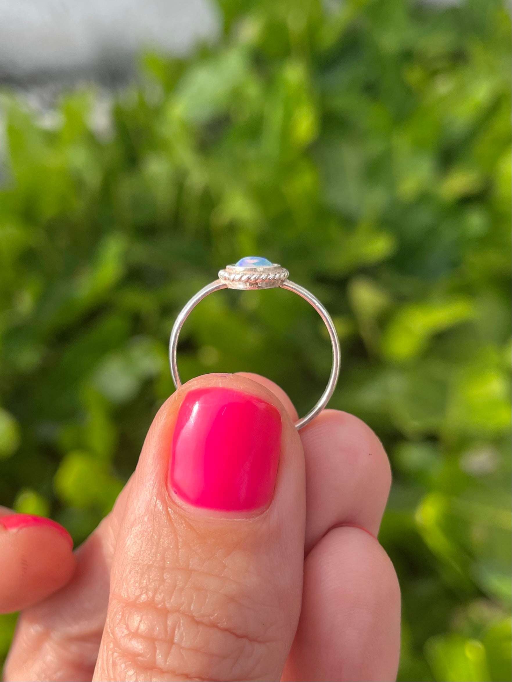 Silver ring with a opal stone held by a hand with pink nail polish against a green leafy background