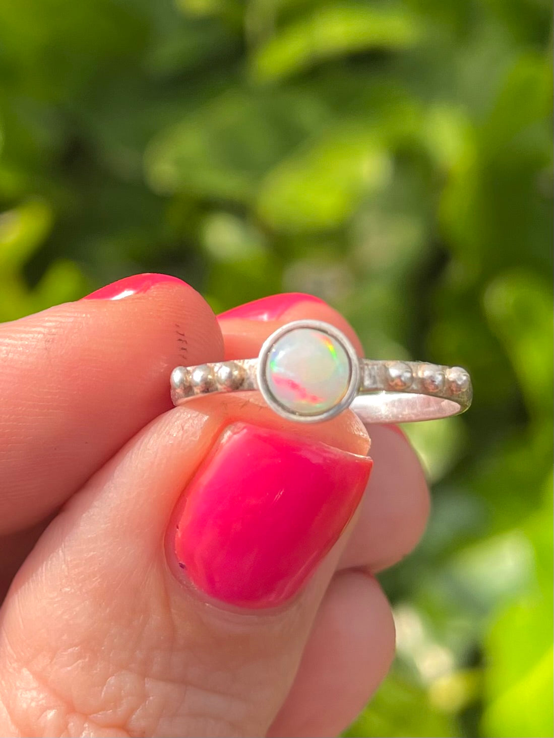 Silver ring with a opal gemstone held by a hand with pink nail polish against leaf background