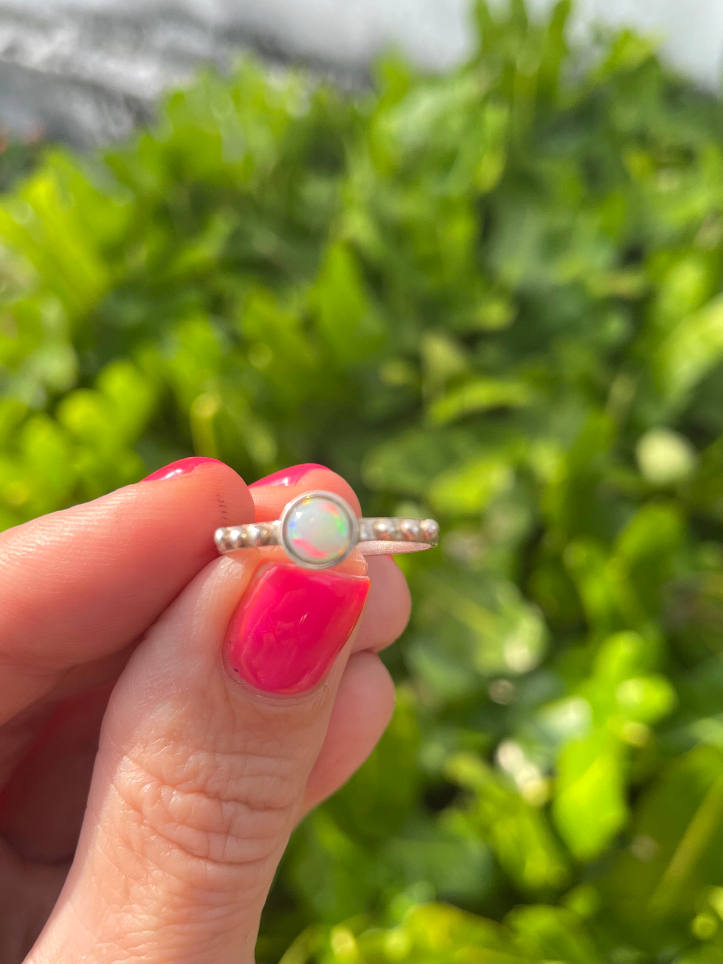 Hand holding a silver ring with an opal stone against a blurred green background 