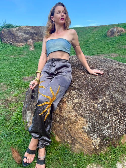 Woman sitting on a rock in a natural setting wearing a denim top and silk upcycled pants with hand embroidered design.
